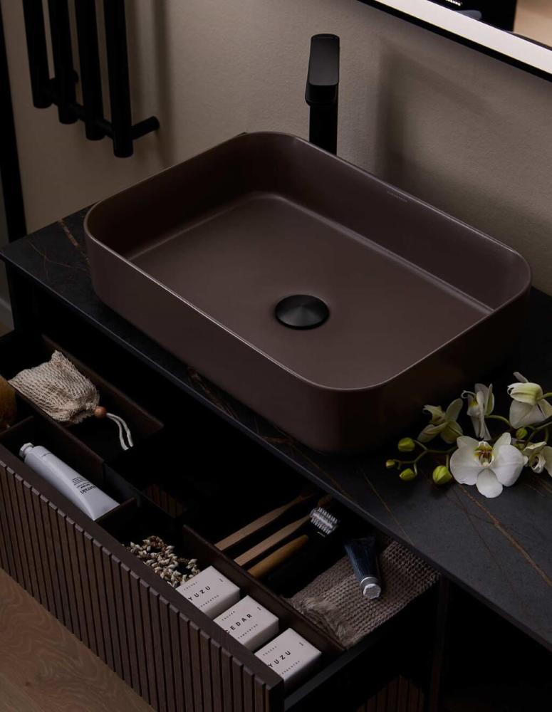 Modern bathroom vanity with a matte brown countertop washbasin, black faucet, and an open drawer neatly organized with soaps, brushes, and toiletries. White orchids rest beside the basin.