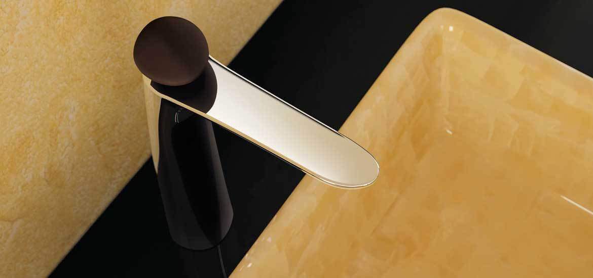 Elegantne kullatoonis valamusegisti musta korpuse ja ümarale juhtnupuga, mille kõrval paikneb kollakas läikiv valamu mustal tööpinnal.