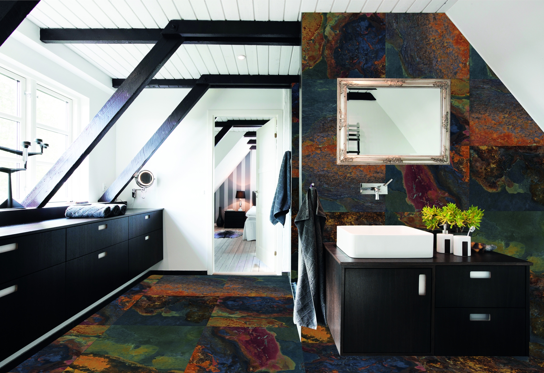 Moodne pööningukorruse vannituba, kus sein ja põrand on kaetud mitmevärviliste kiltkiviplaatidega. Tumeda puiduga vannitoakapp valge valamuga seisab seina ääres, mille kohal on dekoreeritud raamiga peegel. Vasakul on suur aken ja mustade taladega kaldlagi.