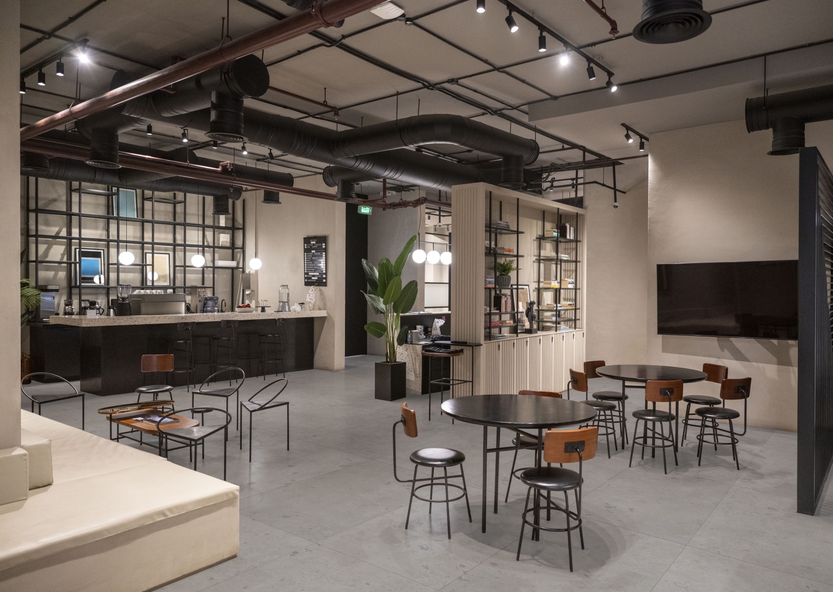 Kaasaegne tööstusliku stiiliga kohvik või ühiskasutatav tööruum, millel on avatud laekonstruktsioon koos mustade torustike ja valgustitega. Ruumi sisustavad mustad ümmargused lauad, metallraamiga toolid, pikk kohvibaar, riiulid raamatute ja taimedega ning suur teler seinal. Põrand on kaetud hallide plaatidega.