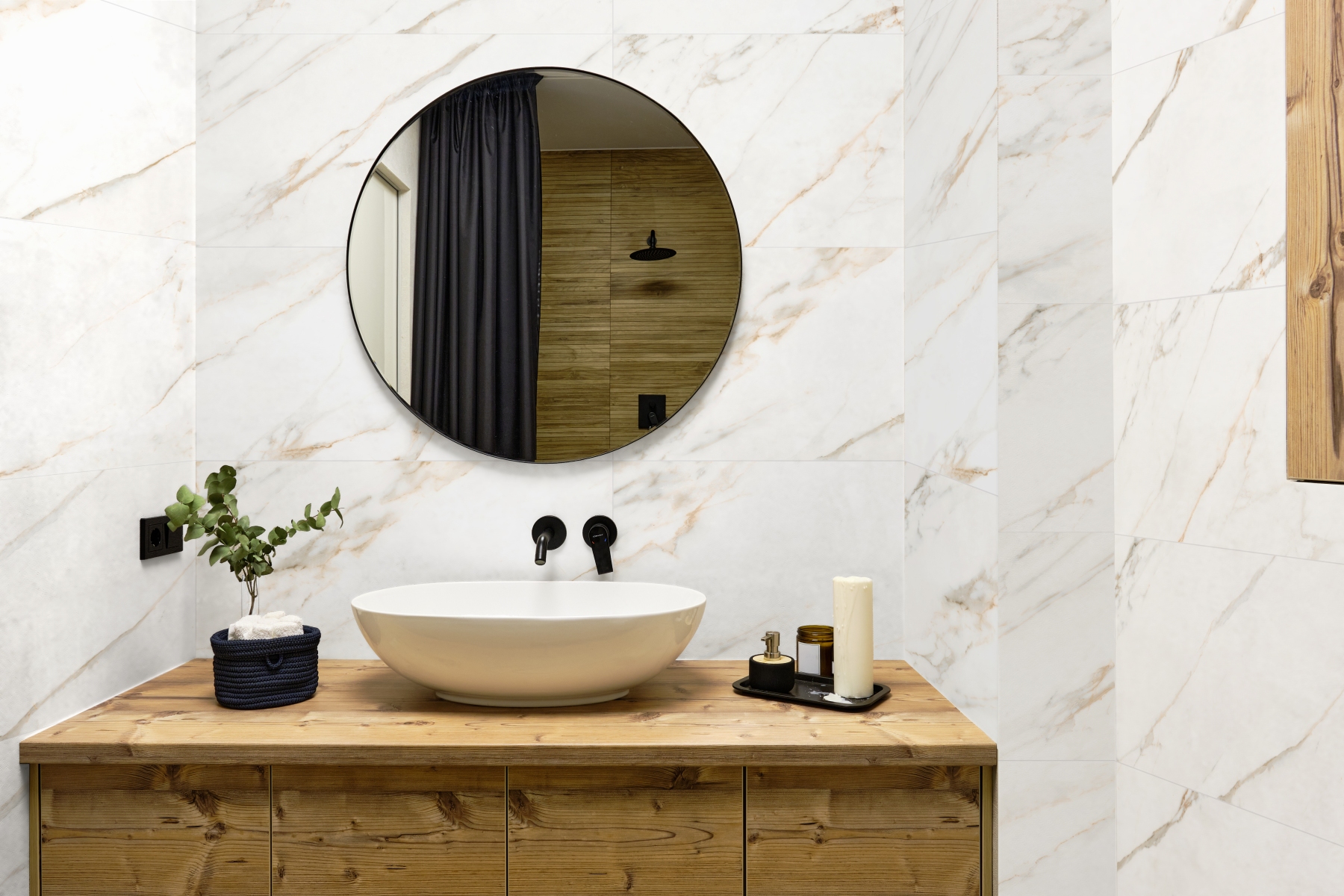 Kaasaegne vannituba heledate marmormustriliste seinaplaatidega. Puidust kapp koos ovaalse valge valamuga ja mustade seinasegistitega loob sooja kontrasti. Ümmargune peegel peegeldab dušinurka, millel on tume kardin ja puidumustriline seinaviimistlus. Dekoratsioonidena on kasutatud rohelist taime, küünalt ja hooldustooteid.