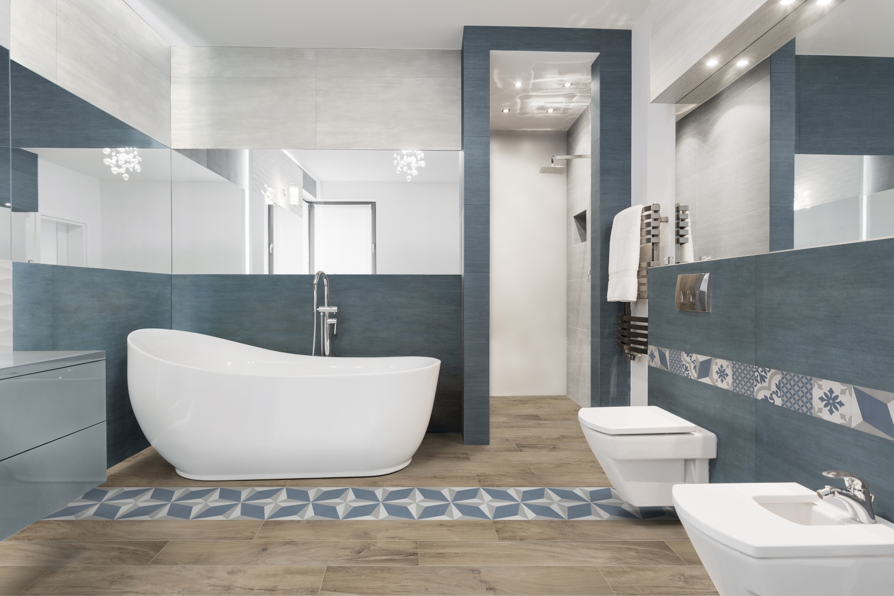 Moodne vannituba, kus on suur vabaduseisuga valge vann, sinakashallide ja heledate plaatidega seinad, puidumustriga põrand ja dekoratiivne geomeetriline aktsentplaatide riba. Paremal on WC-pott ja bidee, taustal asub dušinurk. Interjöör on hele, puhas ja kaasaegne.