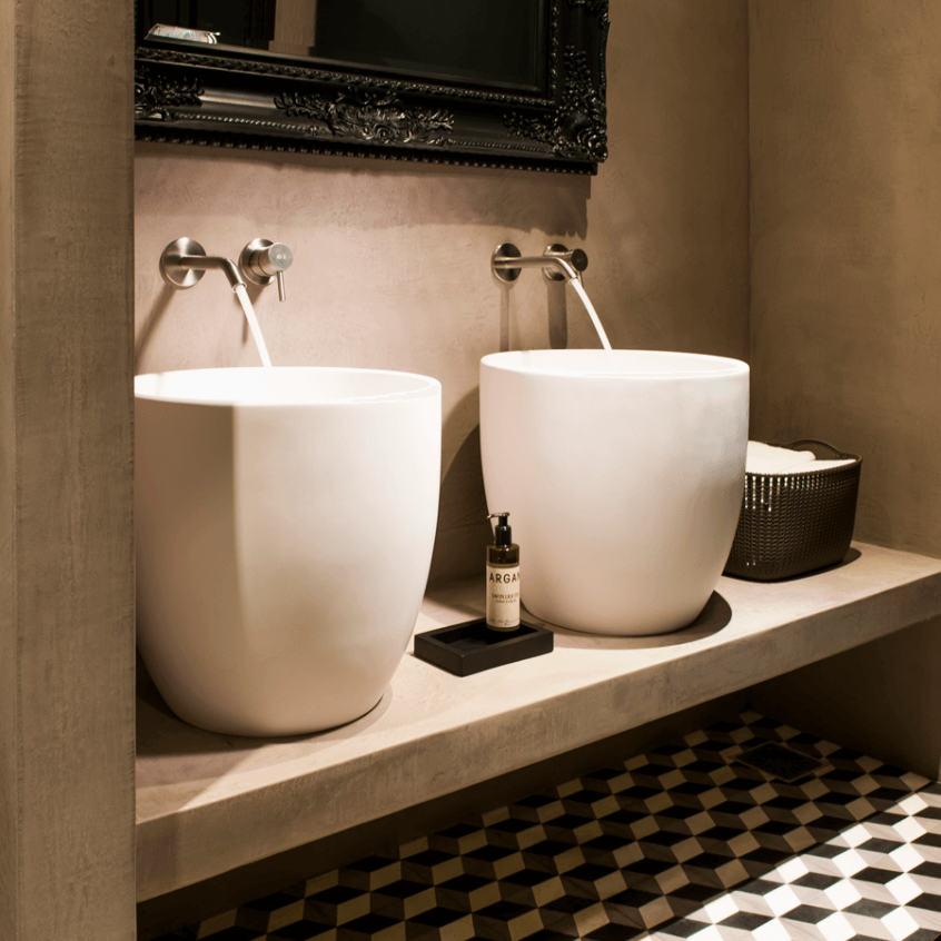 Kaasaegsed mattvalged põrandal seisvad valamud koos seinale paigaldatud segistitega, mida täiendab must dekoratiivne peegel ja ruumiline mustriga põrandakate.