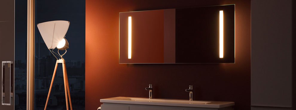 Tume vannituba valgustatud peegli, topeltkraanikausi ja kolmjalgse põrandalambiga.