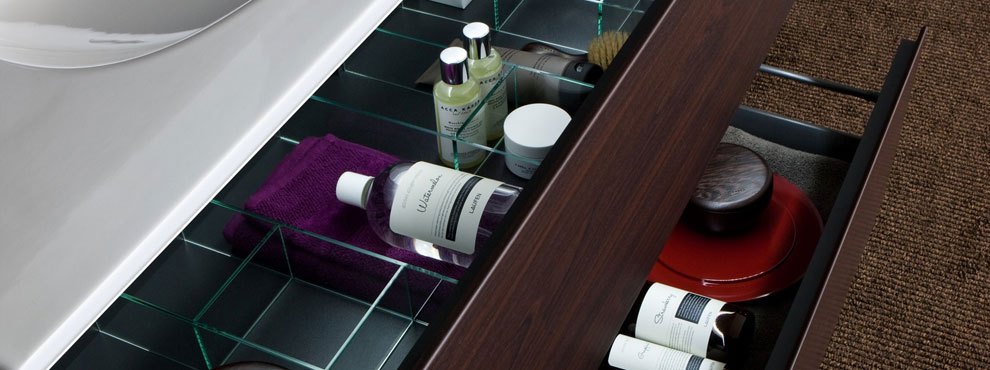 Tume puitviimistlusega vannitoakapp avatud sahtlitega, milles on hooldustooted, pintsel, rätik ja punased karbid.