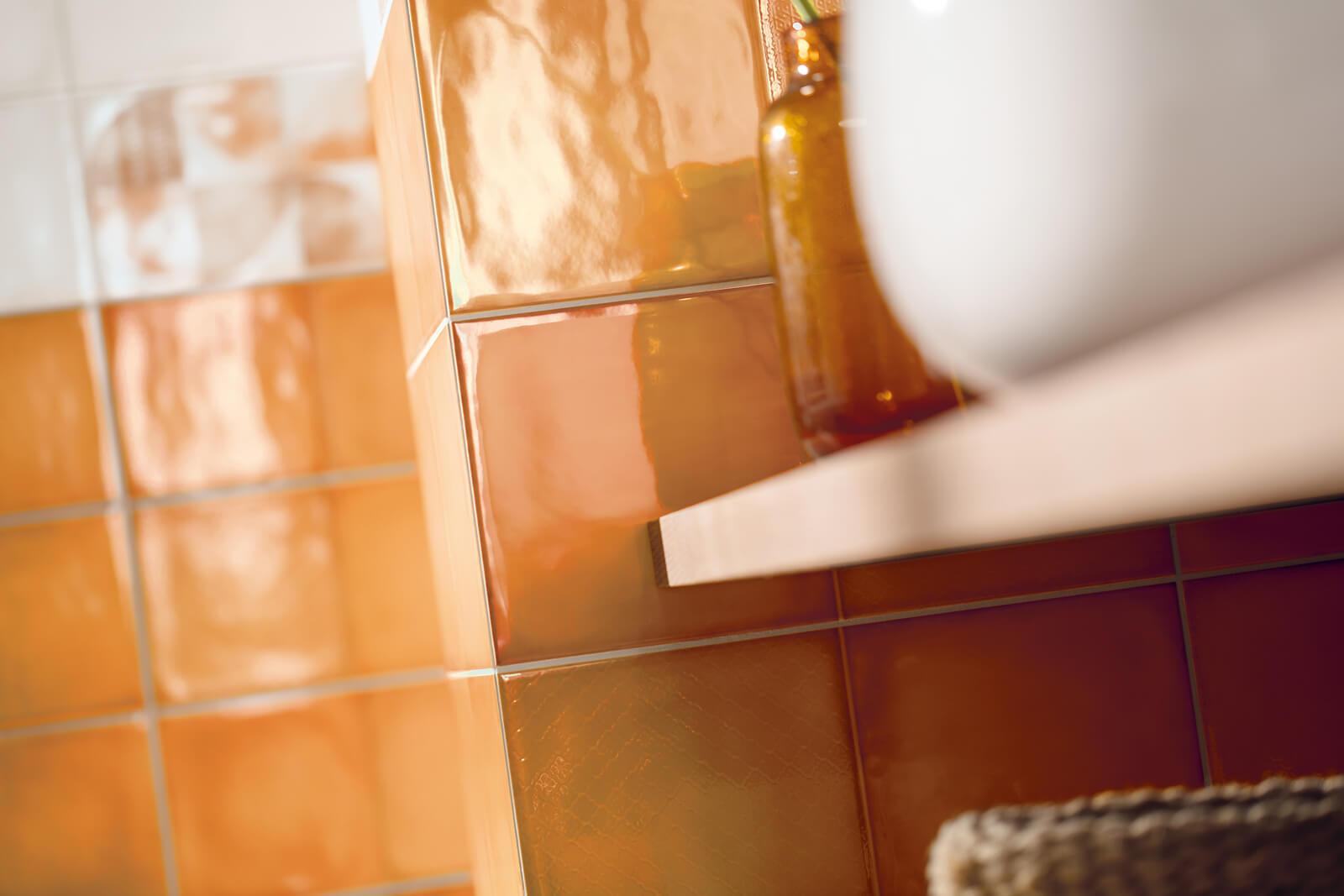 Läikivad oranžid seinaplaadid detailvaates, kõrval pruun klaaspudel ja valge anum riiulil.