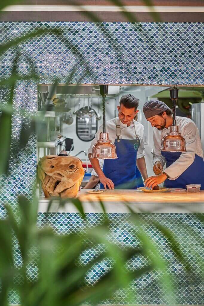 Restorani k&ouml;&ouml;k vaatega l&auml;bi akna, taustal sinise kalasoomust mustriga mosaiikplaat ja kokad t&ouml;&ouml;hoos.