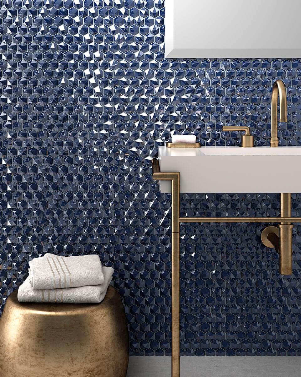 Tumesinistest kuusnurksetest klaasmosaiikidest vannitoa sein, mille ees on kuldsete detailidega valge kraanikauss ja pronksitooni &uuml;mmargune taburet valgete r&auml;tikutega.